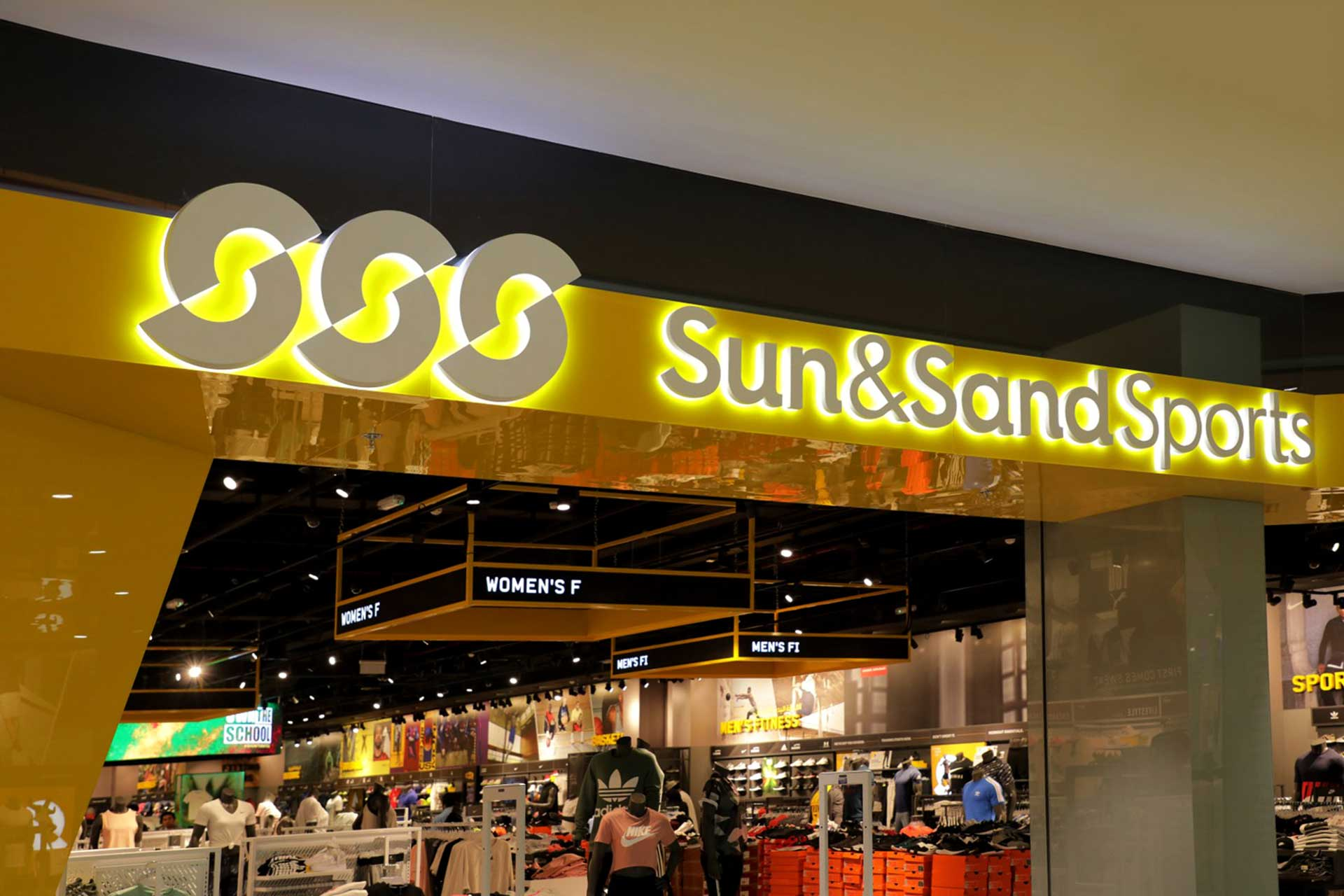 A Sun & Sand Sports store with a bright yellow and black illuminated sign. The store interior is visible, showcasing sports apparel, shoes, and fitness equipment.