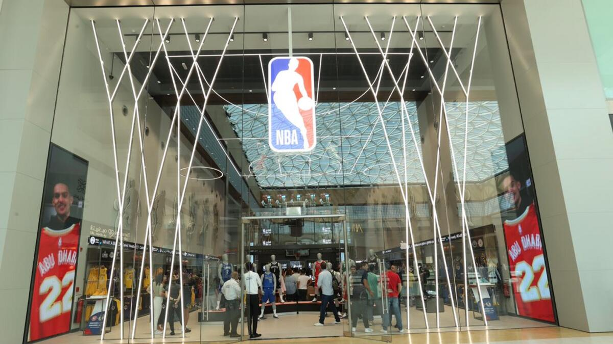 The entrance of an NBA store in a mall, featuring a modern glass facade with crisscross LED lighting. Inside, shoppers are browsing basketball merchandise, jerseys, and sports gear.