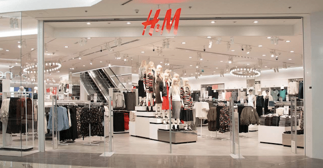 A modern H&M store in a shopping mall with a bright, open layout. The entrance displays mannequins dressed in stylish outfits, with racks of clothing inside.