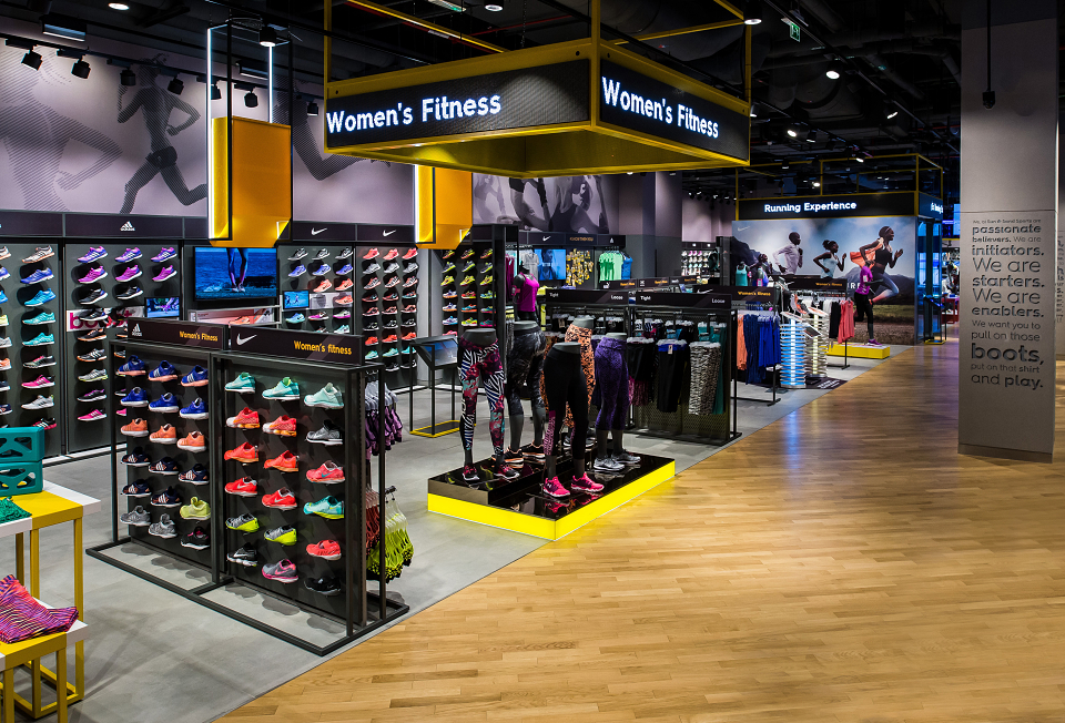 A vibrant women's fitness section in a sports store, featuring colorful athletic shoes, mannequins in workout gear, and a sign reading 'Women's Fitness.' The background includes apparel and running experience sections