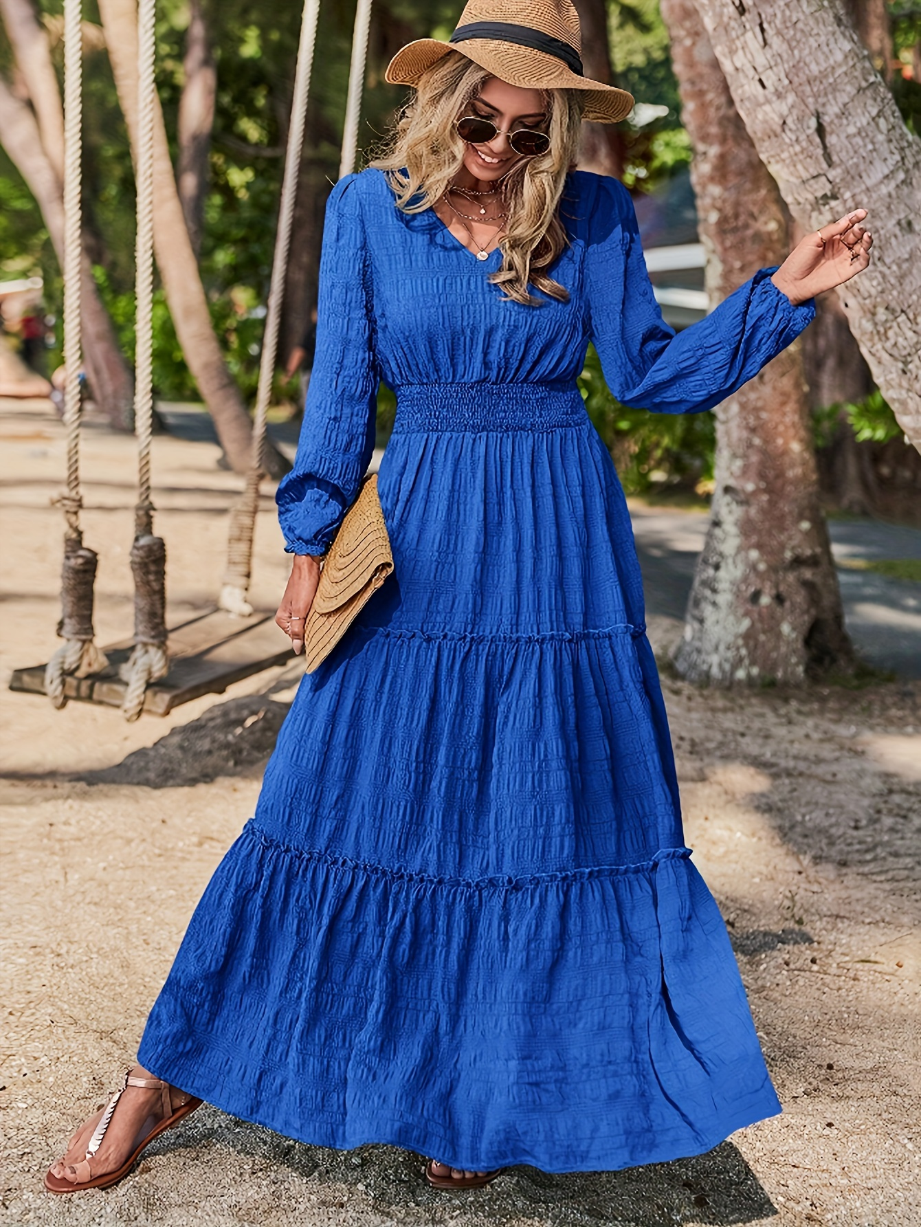 A stylish woman wearing a flowing blue maxi dress, accessorized with a straw hat, sunglasses, and a clutch. She poses outdoors near swings, with trees and sandy ground in the background.