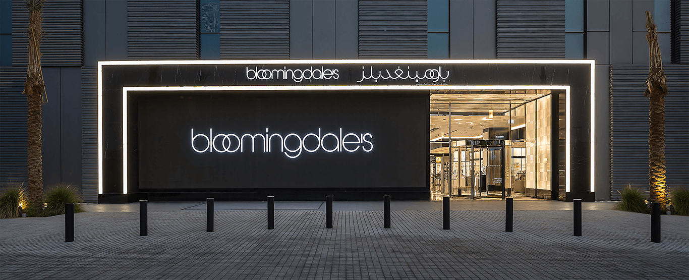 A modern Bloomingdale’s store entrance with a sleek black and illuminated exterior. The signage displays 'bloomingdale's' in both English and Arabic, with glass doors leading into a luxurious retail space.